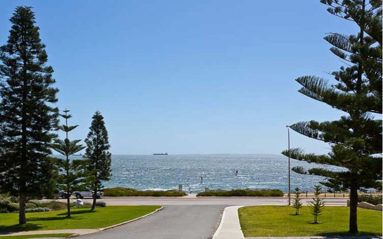 A picture down a clean street which looks out to the ocean showing pine trees in the foreground.