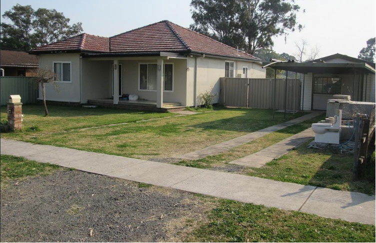 Photo of a bare-bones 3 bedroom weatherboard house and carport with junk in the front yard. A toilet is samongst the junk.