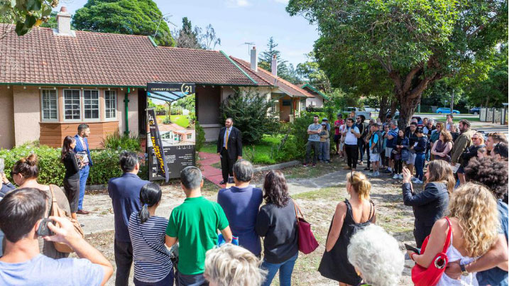 Picture of a house from the curb with a for-sale sign surrounded by an auctioneer and many people watching
